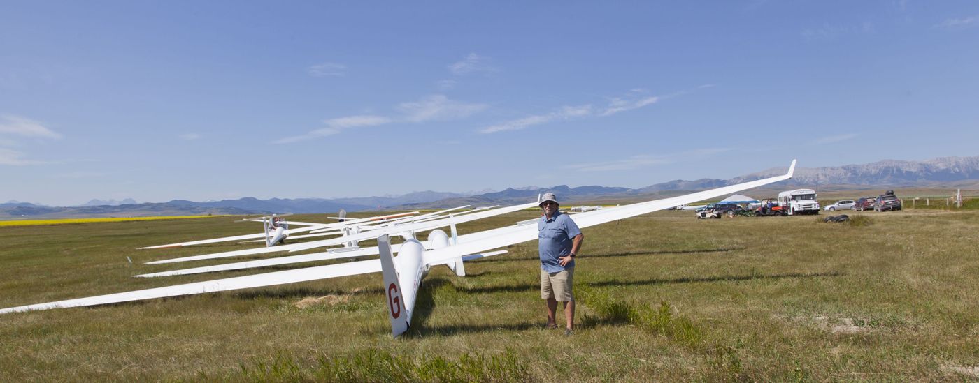 Bashar standing by a line of gliders on a grass airfield with mountains behind